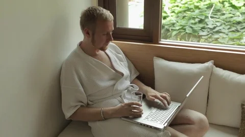 Man in a robe working behind a laptop and drinking water on a sofa by the window Stock-Footage 85278674