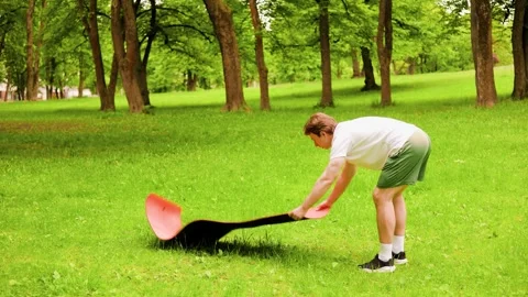 Man rolling out a yoga mat on green grass in a park, preparing for morning yoga  Stock Footage 337493302
