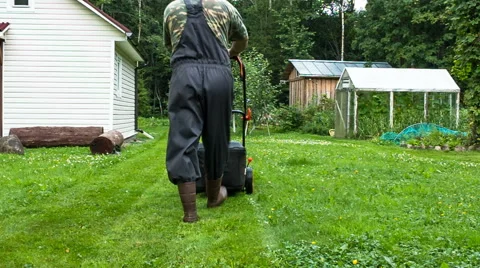 A man in rubber boots walks away from the camera with a lawn mower Stock Footage 65566566