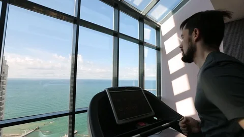 Man running on a treadmill at the panoramic window overlooking the sea Video stock 92528652
