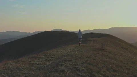 A man runs down the top of a hill against a mountain ridge and a sunset-lit sky Stock Footage 131010310