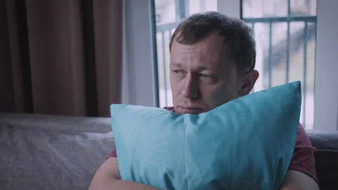 A man with a sad face sits on the sofa, hugging a pillow, with a window in the Stock Footage 314016867