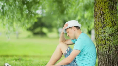 Man is sad while sitting under a tree in a city park, blurred background Stock Footage 148338864