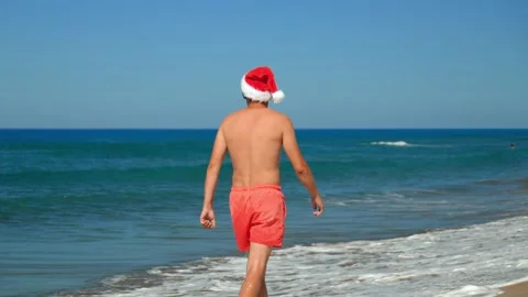 Man in Santa Claus hat walks along sea on background of mountains. Video stock 253391289
