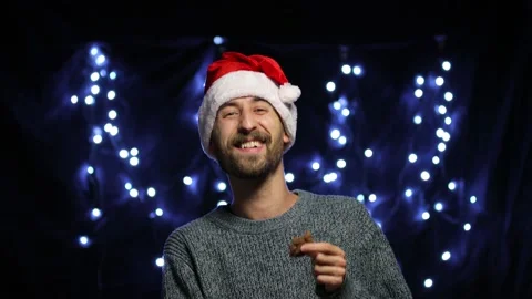 A man in a Santa hat on a bokeh background eats a chocolate cookie Stock Footage 142125399