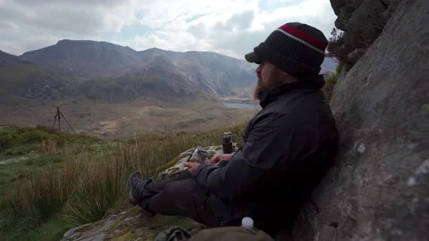 A man sat down relaxing while eating crips in the mountains 動画素材 229146993