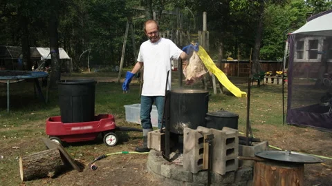 Man scalding butchered broiler using large pot with heated water over fire pit Stock Footage 250733576