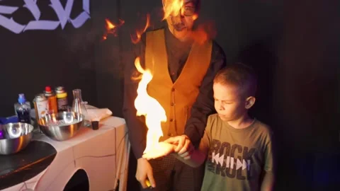 Man with scary makeup sets fire to a child hand against a black wall background Video stock 161990769