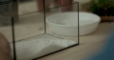 Man scooping white pebbles into clear glass container to shape terrarium base Stock Footage 232080696