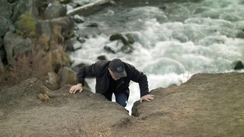 Man scrambles up a steep cliff high above a rapids Stock Footage 99598628