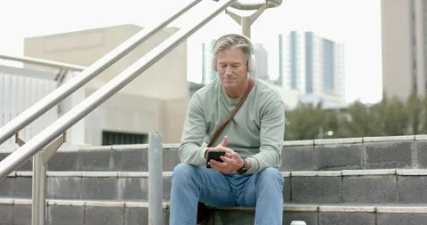 Man scrolling smartphone on plaza steps, hearing headphone audio, glancing at Stock Footage 341851696