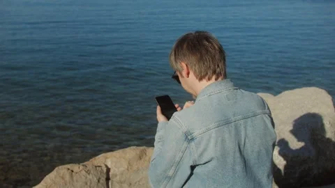 A man by the sea, using smartphone, enjoying the view and relaxing in the sunny Stock Footage 277418340