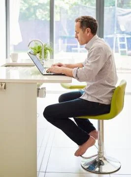A man seated on a kitchen chair at a counter, using a laptop computer. 스톡 사진