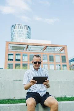 Man seated on steps using a tablet in a modern urban setting during a sunny d Stock Photos