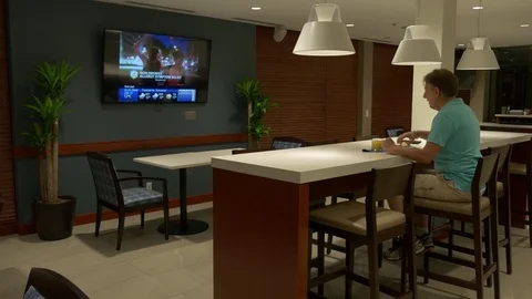 A man is seating on high stool in cafeteria, eating and watching TV. Stockbeeldmateriaal 77084925