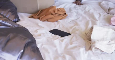 Man seeing face-up black phone on white bed, reaching across, touching sliding Stock Footage 336175089