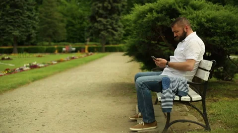Man sends sms and talking for cell phone, sitting on a bench at the park Stock Footage 64283078