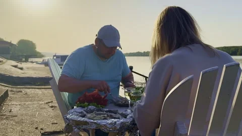 Man Serving Grilled Fish at Riverside Picnic Table 스톡 동영상 334134790