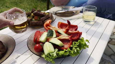 Man Serving Grilled Sausages and Vegetables on Outdoor Table by River, Summer Pi Video stock 333922553