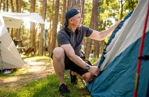 Man sets up blue tent in pine forest during bright daytime Stock Photos