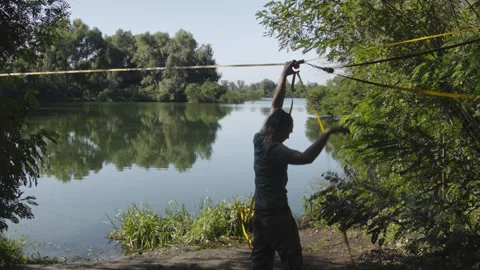 Man sets slackline between trees above the river. 스톡 동영상 140812235