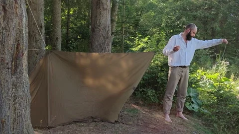 Man setting up camping tarp shelter between trees in mountain forest Stock Footage 344894036