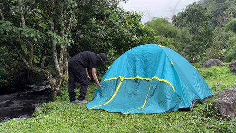 Man Setting Up Camping Tent by Stream in Forest, Misty Mountains, Wide Shot Stock-Footage 324502236