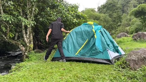 Man Setting Up Green Camping Tent by Stream in Forest Video stock 323676186