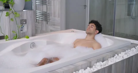Man settling into bubble bath, reclining on headrest in modern bathroom, Stock Footage 348223614