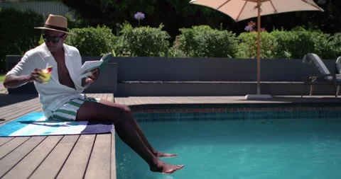 Man is settling at pool edge, reading paperback, sipping drink, dipping feet in Stock Footage 335131548