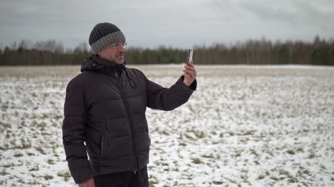 Man in shabby winter jacket having an online video chat standing in a rural Video stock 124064448