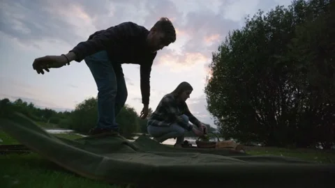 Man Shakes Out Blanket While Woman Prepares Dark Sunset Lakeside Picnic Vidéo 209151488