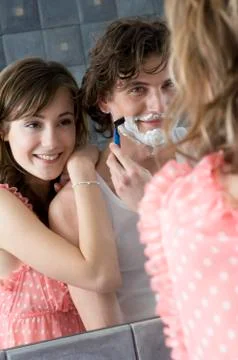 Man shaving in front of bathroom mirror, smiling woman resting on his shoulder Stock Photos