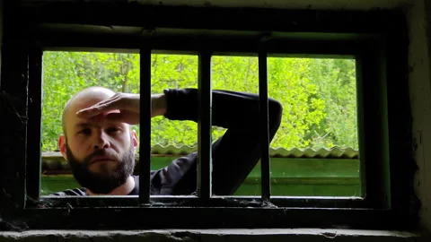 Man shielding eyes to peer through a barred window of old house Video stock 336971281