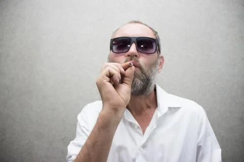 A man in a shirt with a beard in glasses smokes a cigarette. Stock Photos