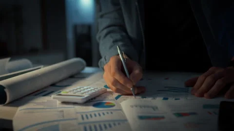 A man in a shirt writes notes with a pen directly on paper charts on a desk. Stock Footage 348317332