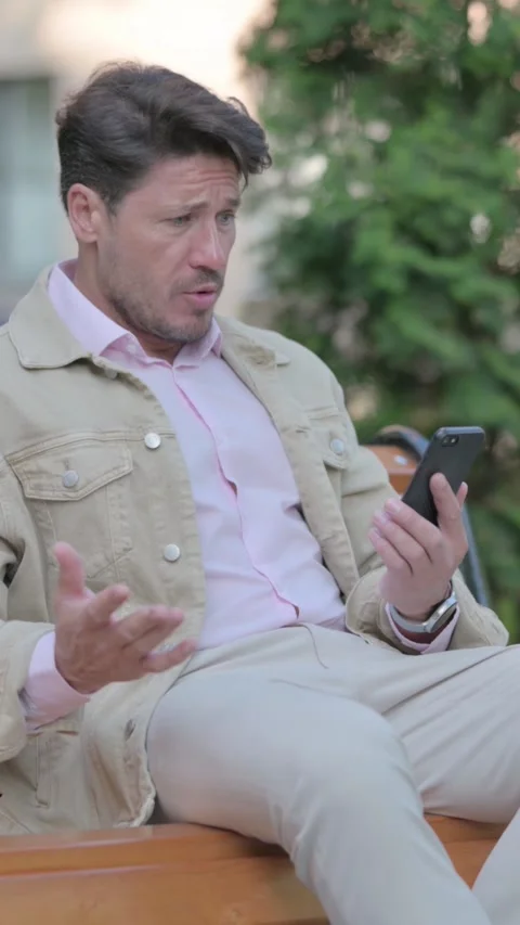 Man Shocked by Loss on Smartphone while Sitting Outdoor on a Bench, vertical Video stock 326267020