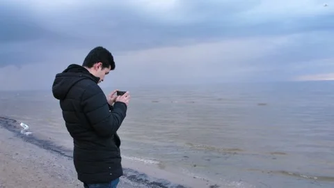 A man by the shore with smartphone on cold, gray day, surrounded by a winter sea Stock Footage 279073686