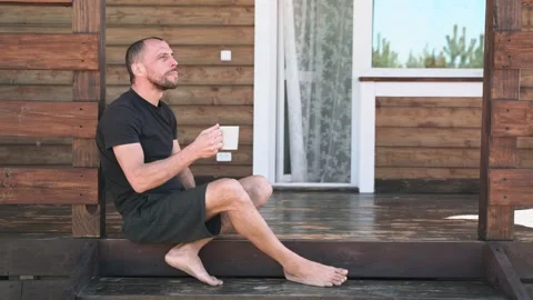 A man in shorts drinks tea sitting on the terrace and enjoys nature The terrace Stock Footage 153972673