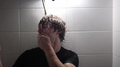 Man in shower covers face with hand as water flows over his head holding smartph Stock Footage 315991963
