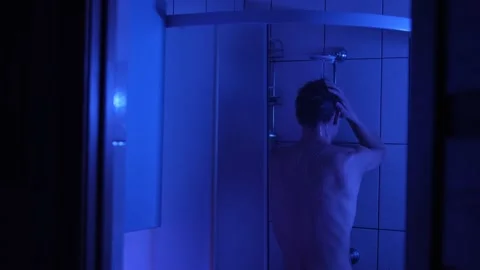 Man in a shower under dim blue light, covering his face in grief, reflecting on Video stock 311452121