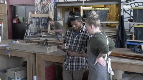 Man showing design on digital tablet to female apprentice in community workshop Stock Footage 220467842