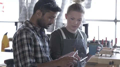 Man showing design on digital tablet to female apprentice in community workshop Stock Footage 220467844