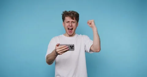 Man shows excitement while playing a game on his phone against a blue backg.. 스톡 동영상 331990856
