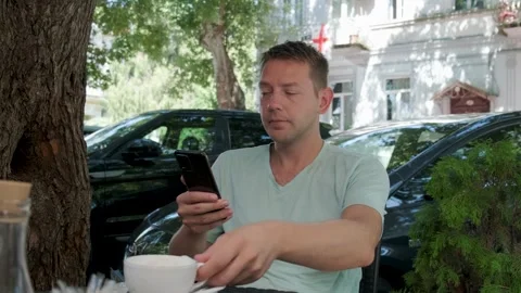 Man in sidewalk cafe using mobile phone and drinking cup of coffee cappuccino Stock Footage 252767704