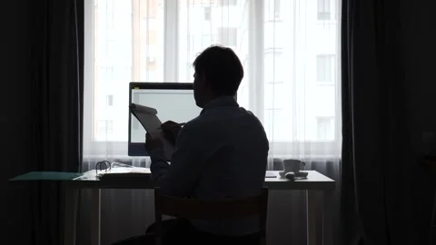 Man in silhouette sitting at desk in front of a window, working on a computer Stock Footage 346251548