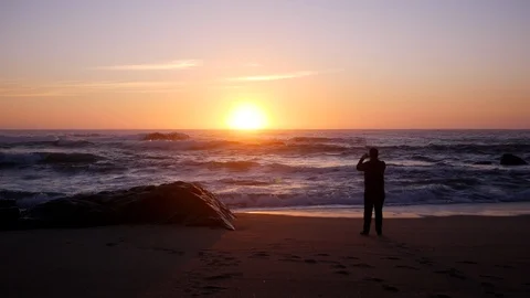 Man in silhouette at sunset on beach, taking photo with cellphone Stock Footage 104683193