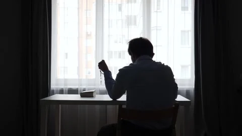 Man in silhouette at a window, seated at a table holding a rosary and praying Stock Footage 345531915