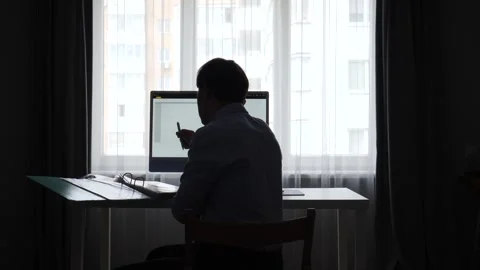 Man silhouette working at a desk, looking at a smartphone while a computer Stock Footage 346251094