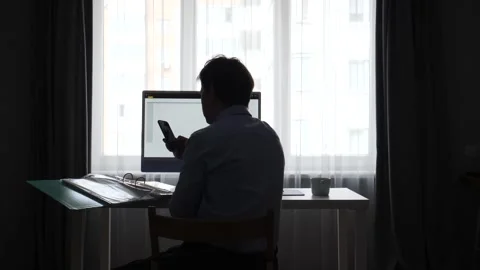 Man silhouette working at a desk by a window, using a computer and smartphone Stock Footage 346251340